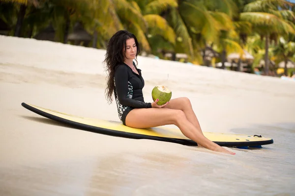 Portret van een mooi surfmeisje met groene kokosnoot uit palmboom zit op geel surfplank. — Stockfoto
