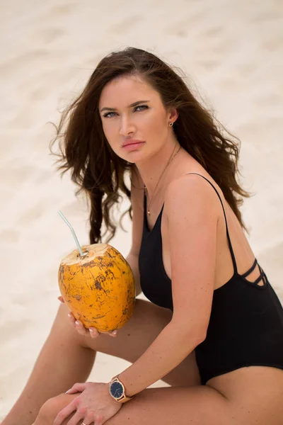 Mujer joven en traje de baño negro con cóctel de coco en la playa . — Foto de Stock