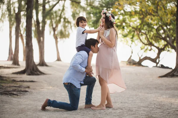 Щаслива вагітна сім'я веселиться в природі. Син на плечах татуся. Батько цілує вагітну дружину в живіт.. — стокове фото