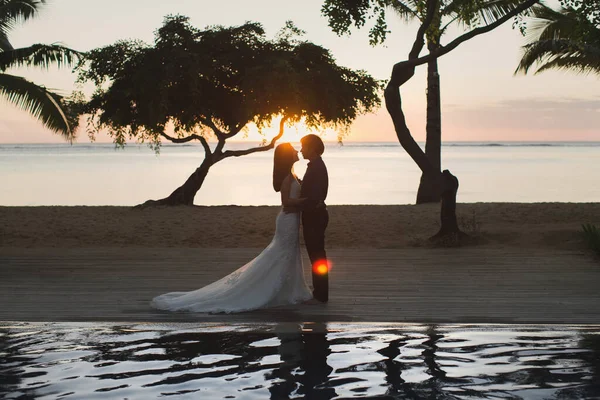 A menyasszony és a vőlegény sziluettjei naplementekor. A tükröződés a medencében. — Stock Fotó
