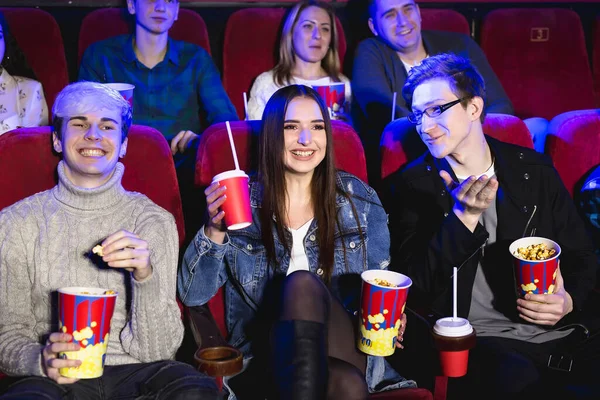 Dois jovens e uma rapariga a ver uma comédia num cinema. Jovens amigos assistem filmes no cinema. Grupo de pessoas no teatro com pipocas e bebidas . — Fotografia de Stock