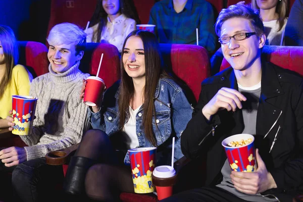 Два молодых парня и девушка смотрят комедию в кинотеатре. Молодые друзья смотрят фильмы в кино. Группа людей в театре с попкорном и напитками . — стоковое фото