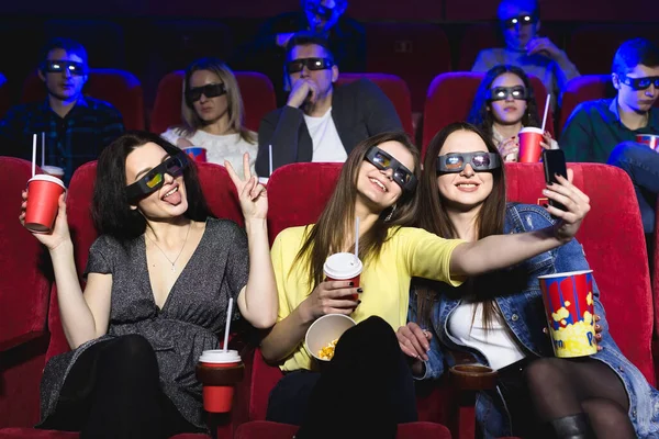 Three beautiful female friends laughing happily making a selfie together during a movie at the cinema. Stock Picture