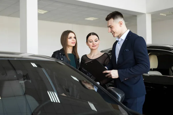 Auto business, car sales - a couple of friends of girls with a car dealer choose a car in a car dealership.