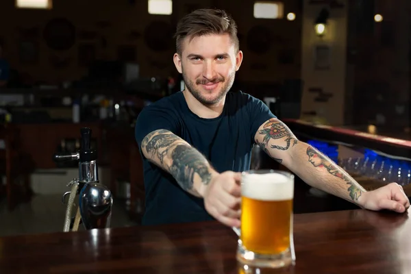 Jóképű fiatal férfi csapos nyújtózkodik üveg sörrel és mosolyog, miközben áll a pultnál. — Stock Fotó