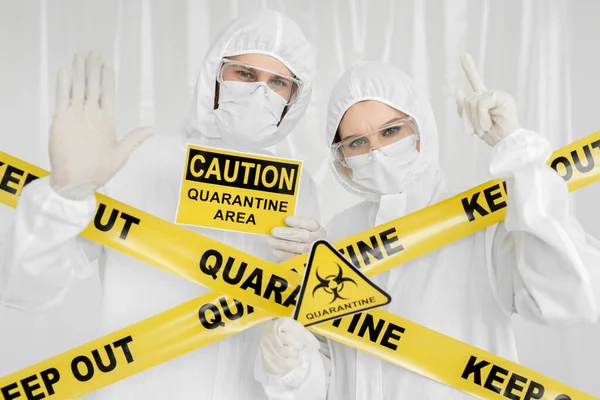 Epidemiologists a man and a woman in protective clothing are in a restricted area with a danger sign. Yellow line Keep Out Quarantine. Entrance is forbidden in quarantine zone. Coronavirus, covid-19. — Stock Photo, Image