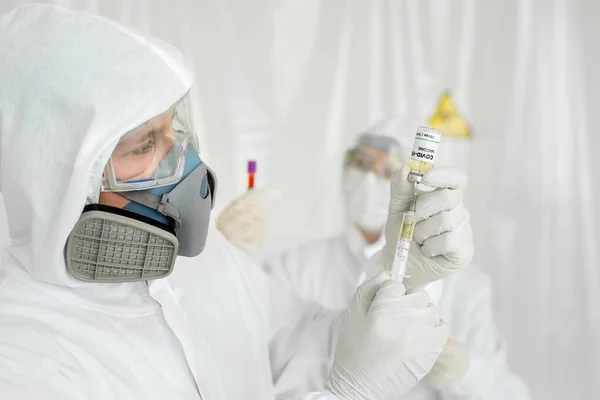 Arzt in Schutzanzug und Maske hält Spritze und Impfstoff COVID-19 in der Hand. Laborwissenschaftler in einem Overall bei der Durchführung eines Forschungsimpfstoffs — Stockfoto