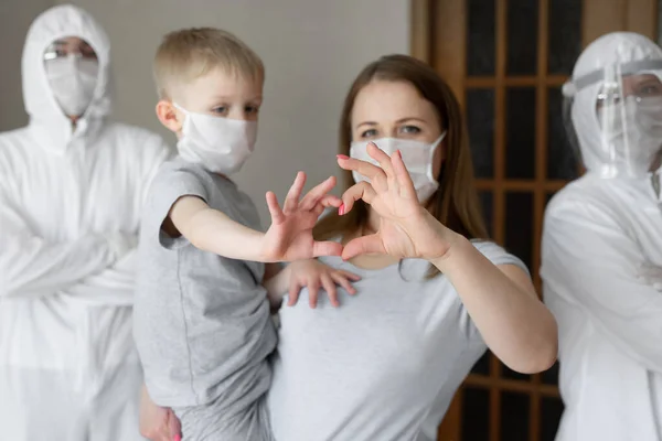 Mutter und Sohn zeigen vor dem Hintergrund von Mitarbeitern in weißen Schutzanzügen während der Coronavirus-Epidemie mit ihren Händen ein Herzzeichen. Covid-19 — Stockfoto