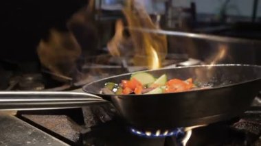 Kişi açık alev üzerinde sebze kızartma tavası sallar