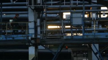Yan görüş işçisi büyük boru hatlarında metal platform boyunca yürüyor ve arka planda parlak güneş ışığı