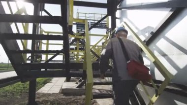 Mavi koruyucu kask içinde closeup çalışan petrol fabrika binası karşı parlak güneş ışığı üzerinde merdivenlerden tırmanıyor