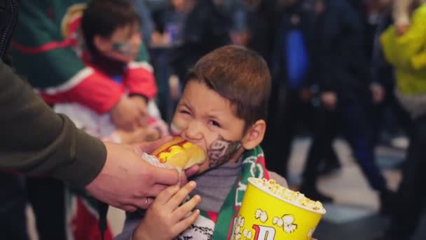 Little boy bites tasty hotdog holding popcorn slow motion — ストック動画