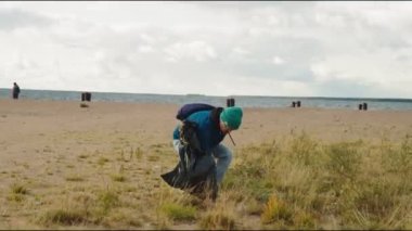 Plastik çantalı genç adam boş kumsalda çöp topluyor.