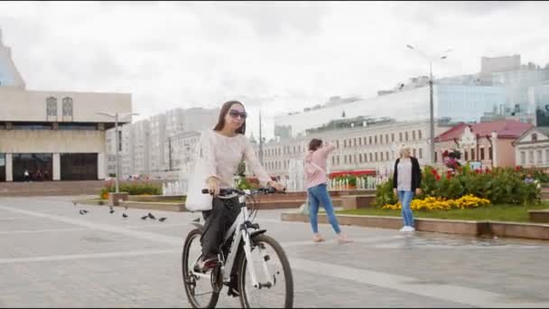 Дівчина в спідниці їде на велосипедах повз фонтани на міській площі. — стокове відео