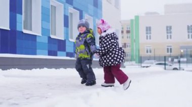 Erkek ve kız karlı anaokulunun bahçesinde el ele yürür.