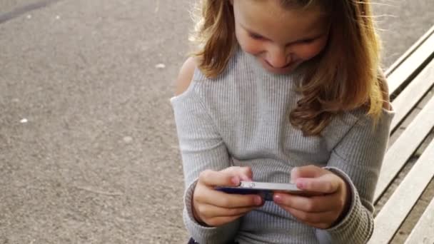 Uma rapariga a falar ao telefone com um amigo. Menina feliz no parque jogando no smartphone — Vídeo de Stock