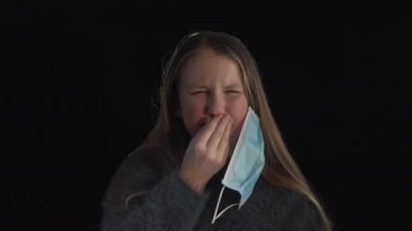 Tatlı hasta kız hapşırır, hasta kız tıbbi maske takar. Siyah arka planda tıbbi maskeli hasta bir kızın portresi. Öksür. Koronavirüs konsepti