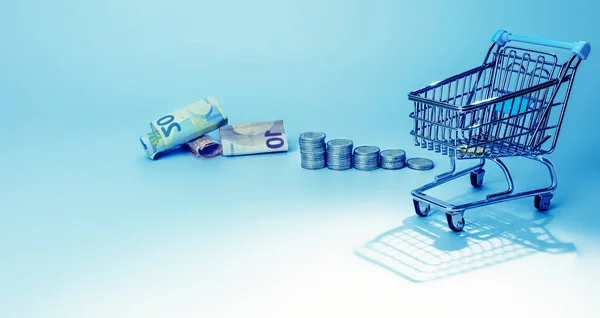 Shopping trolley  for supermarket with money and coins on gradient blue background.The conception of how to cost effective spend money while shopping.