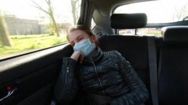 Ağız maskesi takmış hasta bir kız arabanın arka koltuğunda oturur ve pencereye bakar.