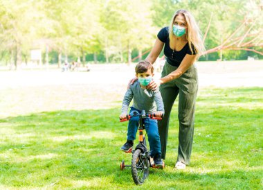 Anne ve oğlu çocuk parkında maskeli. İnsanlar salgın veya grip salgını sırasında koruyucu bir tıbbi maske takarlar. Kişisel koruma ekipmanları. Anne oğluna antiseptik verir. Seçici odak