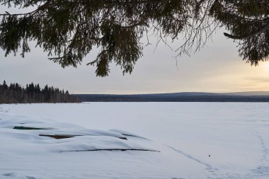 Şafak vakti karla kaplı göl ve kıyıda tekneler. Dağlarda karla kaplı köknar ağaçları olan kış ormanı..