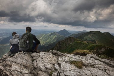 Genç çift dağlarda yürüyüş yapıyor, kayaların üzerinde oturuyor ve manzaraya bakıyor.