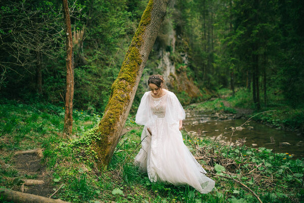 The bride holds her dress and walks through the woods along the river