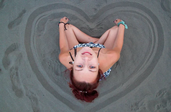 Fille rousse assise dans le sable sur la plage, coeur dessiné dans le sable nom — Photo