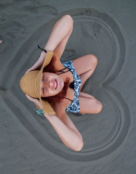 Une rousse avec un chapeau sur la tête est assise dans le sable sur le be — Photo