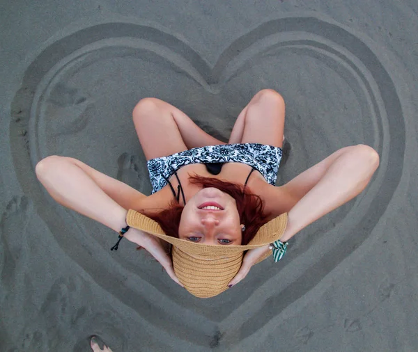 Een roodharige meisje met een hoed op haar hoofd zit in het zand op de worden — Stockfoto