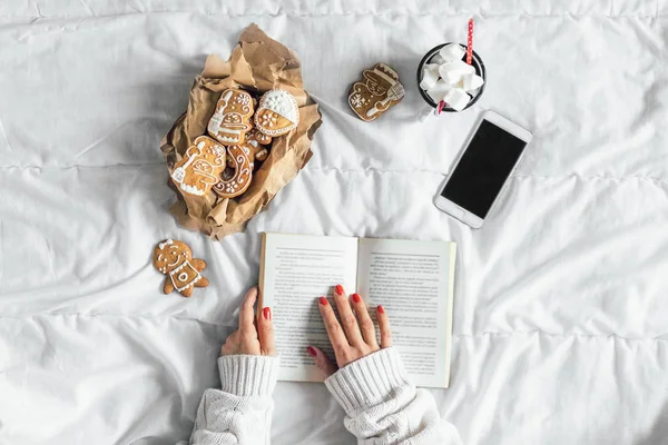 woman reading book. Top view. Smartphone, hot chocolate  with marshmallows and cookies on white sheet