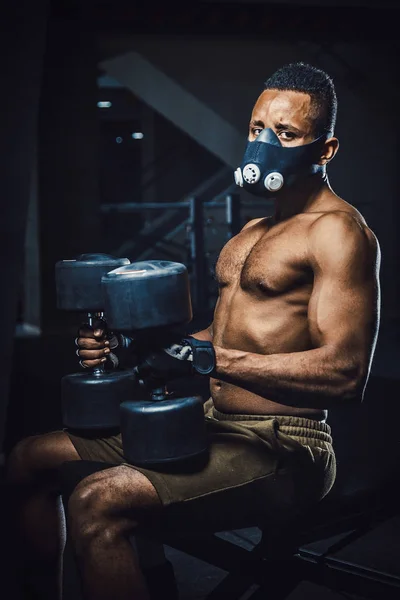 oturma ve bekleyen ve aynanın önünde halter kaldırma önce kameraya bakarak maskeli Afro-Amerikan atletik adam. zenci dumbbells ile spor salonunda