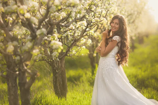 在鲜花的美丽女子的画像。新娘在象牙色婚纱，长长的卷发在夏天开花树木的花园里散步 — 图库照片