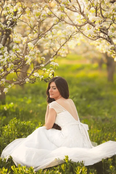 Porträt einer schönen Frau in Blumen. die Braut im elfenbeinfarbenen Brautkleid mit langen lockigen Haaren, die in Gärten mit sommerblühenden Bäumen spaziert — Stockfoto