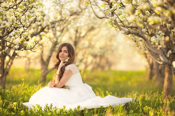 Porträt einer schönen Frau in Blumen. die Braut im elfenbeinfarbenen Brautkleid mit langen lockigen Haaren, die in Gärten mit sommerblühenden Bäumen spaziert — Stockfoto