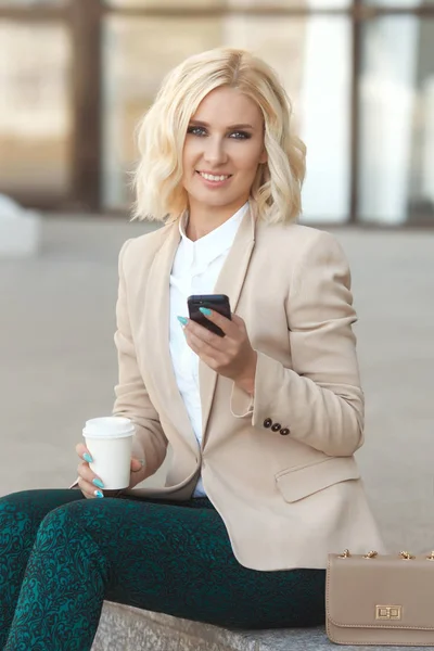 Hermosa mujer de negocios en ropa de moda sonriendo, sosteniendo el teléfono móvil al aire libre y mirando a la cámara —  Fotos de Stock