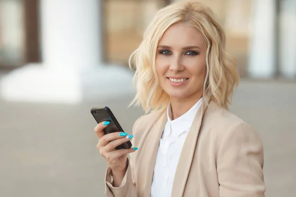 Hermosa mujer de negocios en ropa de moda sonriendo, sosteniendo el teléfono móvil al aire libre y mirando a la cámara —  Fotos de Stock