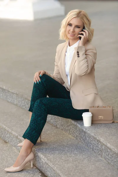 Hermosa mujer de negocios en ropa de moda sonriendo, sosteniendo el teléfono móvil al aire libre y mirando a la cámara —  Fotos de Stock