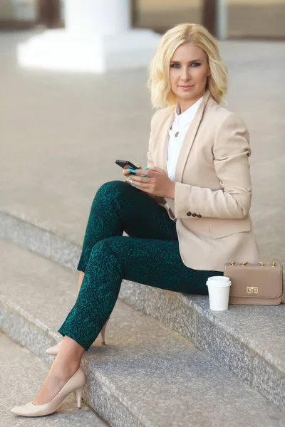 Mooie zakenvrouw in mode kleding glimlachen, het bezit van mobiele telefoon buiten en het kijken naar camera — Stockfoto