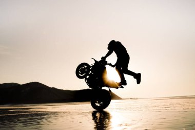 stunt rider riding on back wheel of motorcycle on ice in the sunset