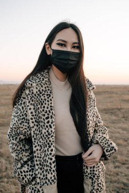 Siyah viral maske takan Asyalı moda kızı ve gün batımında bir tarlada yürüyen leopar desenli ceket.