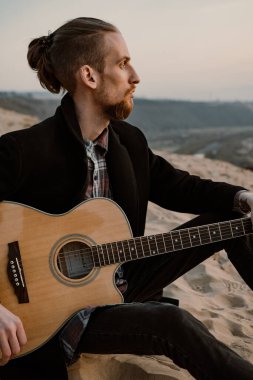 Yakışıklı sakallı adam gün batımında gitarıyla kum tepesinde oturuyor.