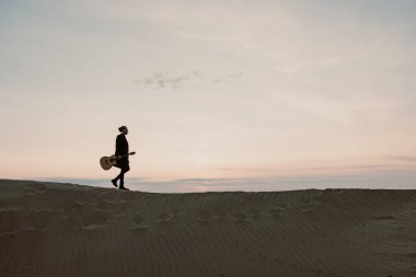 Uzun saçlı, gitarlı bir adam Sibirya 'da gün batımında kumlarda yürüyor.