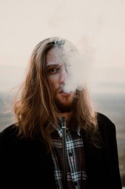 Uzun saçlı sakallı hippi gün batımında pipo içiyor.