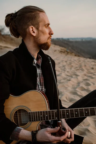 Uzun saçlı sakallı hippi gün batımında tepede gitar çalıyor.