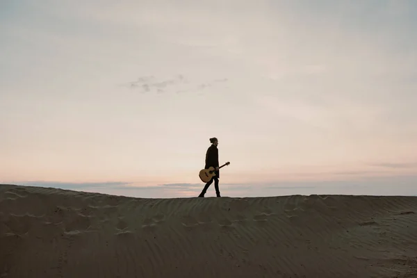Uzun saçlı, gitarlı bir adam Sibirya 'da gün batımında kumlarda yürüyor.