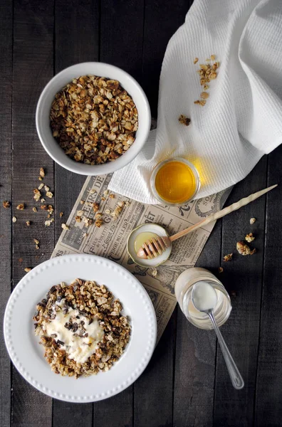 Homemade oatmeal granola with peanuts, yogurt and honey on white plates on dark table, sunny morning