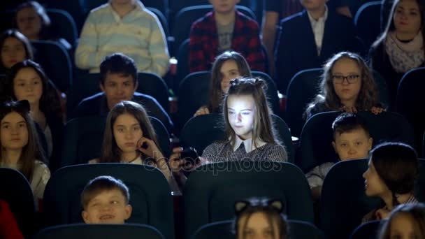 Lilla vackra flicka ser på sig själv i biografen hall. Många barn sitter. — Stockvideo