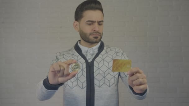 Egy fiatal férfi-érme bitcoin és banc kártyával. Az ember úgy néz ki, és hasonlítja össze. Fényképezőgép, 4 k. — Stock videók