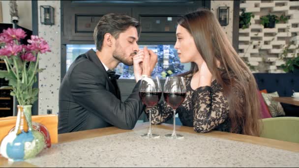 Joven hombre besa la mano de una mujer bonita, en la mesa dos copas de vino. Hay una tabla de snowboard en el suelo . — Vídeo de stock